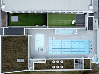 An aerial view of a modern rooftop facility featuring a large swimming pool with multiple lanes, adjacent to a smaller pool or water feature. The rooftop has landscaped grass areas with what appears to be a tennis court setup on one side, and sections covered with foliage, possibly for insulation or eco-friendly purposes. The design is geometric, with clean lines and a mix of concrete and greenery.
