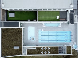 An aerial view of a modern rooftop facility featuring a large swimming pool with multiple lanes, adjacent to a smaller pool or water feature. The rooftop has landscaped grass areas with what appears to be a tennis court setup on one side, and sections covered with foliage, possibly for insulation or eco-friendly purposes. The design is geometric, with clean lines and a mix of concrete and greenery.