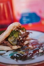 A plate with an intricately designed border holds a sandwich-like dish. The dish consists of a soft, round bun filled with shredded meat, cucumber slices, herbs, and garnished with chopped nuts.