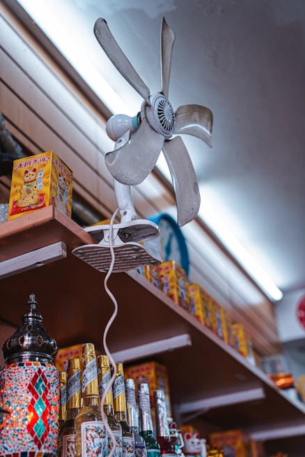 A vintage electric fan is mounted on a high shelf with its blades covered in dust. Below the fan, shelves are stocked with various items, including colorful mosaic lamps and bottled drinks. The shelves also hold several boxes featuring a waving cat design, commonly known as a 'maneki-neko'. A fluorescent light illuminates the scene from above, casting a soft glow.