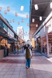 A bustling outdoor shopping area featuring modern architecture with glass-fronted stores on either side of a tiled pedestrian walkway. Overhead, strings of white lanterns create a festive atmosphere. People are walking, some stopping to look at storefronts, while a few are engaged in conversation. The sky is clear with a few wispy clouds, promoting a bright and vibrant ambiance.