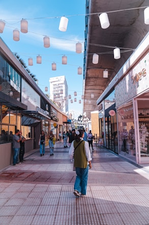 A bustling outdoor shopping area featuring modern architecture with glass-fronted stores on either side of a tiled pedestrian walkway. Overhead, strings of white lanterns create a festive atmosphere. People are walking, some stopping to look at storefronts, while a few are engaged in conversation. The sky is clear with a few wispy clouds, promoting a bright and vibrant ambiance.