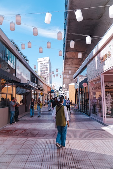 A bustling outdoor shopping area featuring modern architecture with glass-fronted stores on either side of a tiled pedestrian walkway. Overhead, strings of white lanterns create a festive atmosphere. People are walking, some stopping to look at storefronts, while a few are engaged in conversation. The sky is clear with a few wispy clouds, promoting a bright and vibrant ambiance.