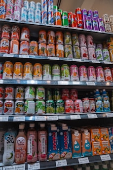 A variety of beverage cans and bottles are neatly organized on supermarket shelves. The selection includes both cans with colorful designs and bottled drinks with unique packaging. Each shelf is labeled with price tags indicating the cost per unit.