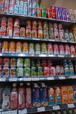 A variety of beverage cans and bottles are neatly organized on supermarket shelves. The selection includes both cans with colorful designs and bottled drinks with unique packaging. Each shelf is labeled with price tags indicating the cost per unit.