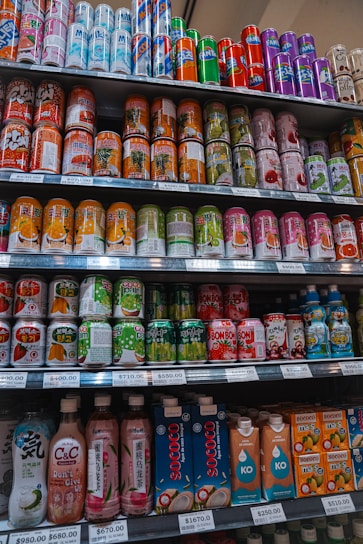 A variety of beverage cans and bottles are neatly organized on supermarket shelves. The selection includes both cans with colorful designs and bottled drinks with unique packaging. Each shelf is labeled with price tags indicating the cost per unit.