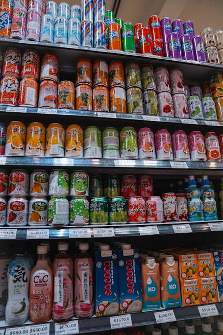 A variety of beverage cans and bottles are neatly organized on supermarket shelves. The selection includes both cans with colorful designs and bottled drinks with unique packaging. Each shelf is labeled with price tags indicating the cost per unit.