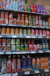 A variety of beverage cans and bottles are neatly organized on supermarket shelves. The selection includes both cans with colorful designs and bottled drinks with unique packaging. Each shelf is labeled with price tags indicating the cost per unit.