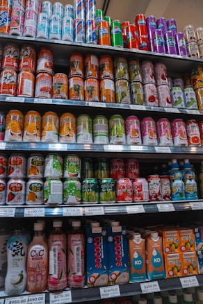 A variety of beverage cans and bottles are neatly organized on supermarket shelves. The selection includes both cans with colorful designs and bottled drinks with unique packaging. Each shelf is labeled with price tags indicating the cost per unit.