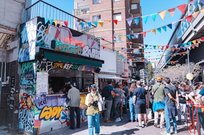 A vibrant street scene with people gathered around a small graffiti-covered kiosk. Colorful triangular banners hang overhead, creating a festive atmosphere. The crowd consists of casually dressed individuals engaging with the vendor and each other on a sunny day. The surrounding buildings add an urban backdrop to the lively setting.