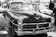 A classic vintage car is prominently displayed in a parking lot, surrounded by other vehicles. The car showcases distinct chrome detailing and a polished exterior. A license plate with the text 'PH 50WAY' is visible at the front. The windows display various signs, hinting at a car show or gathering.