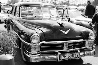 A classic vintage car is prominently displayed in a parking lot, surrounded by other vehicles. The car showcases distinct chrome detailing and a polished exterior. A license plate with the text 'PH 50WAY' is visible at the front. The windows display various signs, hinting at a car show or gathering.