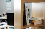 A close-up view of a wooden bookshelf holding three books. The titles visible are 'Berlin', 'Europe and the Sea', and 'American English'. In the background, a blurred office setting with desks, a computer monitor, and some office supplies can be seen.