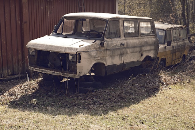 Two old, abandoned vans are positioned next to a wooden building. The front van is noticeably rusted and missing parts like the front grille and headlights. Both vehicles appear dilapidated, surrounded by dry grass and weeds. The scene suggests long-term neglect and abandonment.