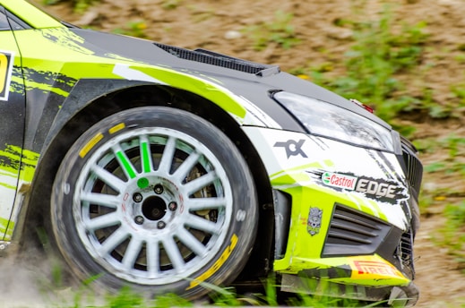 A close-up of a race car's front section during a rally, showcasing its aerodynamic design, green and black livery, visible branding, and detailed alloy wheel spinning, highlighted with sponsor decals.