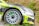 A close-up of a race car's front section during a rally, showcasing its aerodynamic design, green and black livery, visible branding, and detailed alloy wheel spinning, highlighted with sponsor decals.