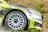 A close-up of a race car's front section during a rally, showcasing its aerodynamic design, green and black livery, visible branding, and detailed alloy wheel spinning, highlighted with sponsor decals.