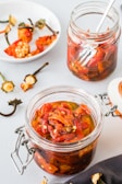 A close-up of a glass jar filled with pickled red peppers in oil, with a spoon in the background. There is another jar of peppers and a plate with pepper stems and seeds scattered around. The scene is set on a light-colored surface with a contrast of dark cloth nearby.