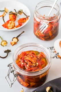 A close-up of a glass jar filled with pickled red peppers in oil, with a spoon in the background. There is another jar of peppers and a plate with pepper stems and seeds scattered around. The scene is set on a light-colored surface with a contrast of dark cloth nearby.