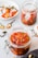 A close-up of a glass jar filled with pickled red peppers in oil, with a spoon in the background. There is another jar of peppers and a plate with pepper stems and seeds scattered around. The scene is set on a light-colored surface with a contrast of dark cloth nearby.