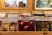 A collection of vinyl records is neatly organized in wooden display crates. The records feature notable musicians and bands such as John Cougar, Bob Dylan, Eric Clapton, and Crosby, Stills, Nash & Young. The background includes a painting with palm trees and a model sailboat, adding a vintage and eclectic atmosphere to the scene.