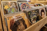 A collection of vinyl records is neatly displayed in wooden storage bins. The covers of the records feature vibrant and diverse artwork, reflecting a variety of musical genres and styles. Some album covers are quite colorful, while others feature prominent portraits of artists.