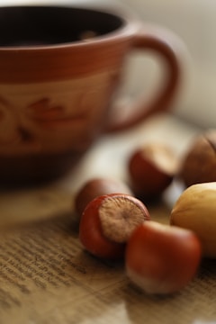 A close-up of several hazelnuts placed on a textured surface, with the focus on the front nut and the rest slightly blurred. A large, brown ceramic mug is visible in the blurry background, contributing to an earthy and warm atmosphere.