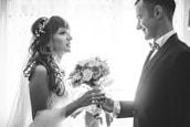 A bride and groom are facing each other, holding hands, in a romantic and intimate moment. The bride is wearing a lace dress with a decorative hairpiece, and she holds a bouquet of flowers. The groom is dressed in a formal suit with a bow tie. The scene is captured in black and white, adding a timeless and classic feel.