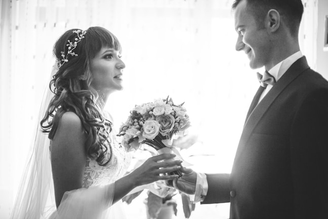 A bride and groom are facing each other, holding hands, in a romantic and intimate moment. The bride is wearing a lace dress with a decorative hairpiece, and she holds a bouquet of flowers. The groom is dressed in a formal suit with a bow tie. The scene is captured in black and white, adding a timeless and classic feel.