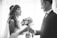 A bride and groom are facing each other, holding hands, in a romantic and intimate moment. The bride is wearing a lace dress with a decorative hairpiece, and she holds a bouquet of flowers. The groom is dressed in a formal suit with a bow tie. The scene is captured in black and white, adding a timeless and classic feel.
