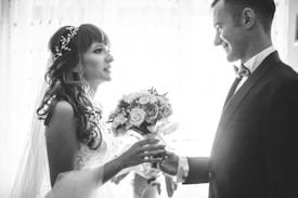 A bride and groom are facing each other, holding hands, in a romantic and intimate moment. The bride is wearing a lace dress with a decorative hairpiece, and she holds a bouquet of flowers. The groom is dressed in a formal suit with a bow tie. The scene is captured in black and white, adding a timeless and classic feel.