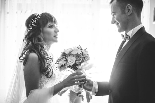 A bride and groom are facing each other, holding hands, in a romantic and intimate moment. The bride is wearing a lace dress with a decorative hairpiece, and she holds a bouquet of flowers. The groom is dressed in a formal suit with a bow tie. The scene is captured in black and white, adding a timeless and classic feel.
