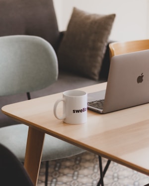 A modern workspace featuring a wooden table with a laptop and a white mug labeled 'swello', accompanied by a few pillows in the background. The setting includes chairs with soft padding, creating a cozy and minimalistic office environment.