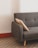 A mid-century modern gray sofa with wooden legs and a beige accent pillow rests against a plain white wall. The floor is decorated with a geometric patterned tile, and there is a reddish-brown wooden trim at the base of the wall.