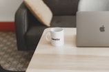 A minimalist setup on a light wooden table with a silver laptop and a white mug displaying the text 'swello'. The background features a dark gray couch with a beige cushion, set against a patterned carpet.
