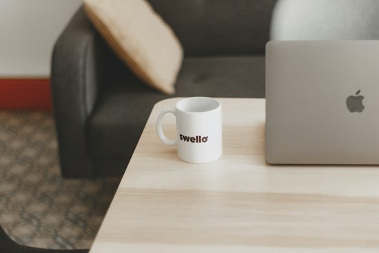 A minimalist setup on a light wooden table with a silver laptop and a white mug displaying the text 'swello'. The background features a dark gray couch with a beige cushion, set against a patterned carpet.