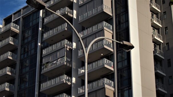 A modern high-rise apartment building with multiple balconies is shown during the daytime. The structure has a sleek, contemporary design with prominent use of glass and steel. The façade is largely in shadow, indicative of the image being taken likely during the late afternoon. Two streetlights are visible in the foreground, adding to the urban atmosphere.