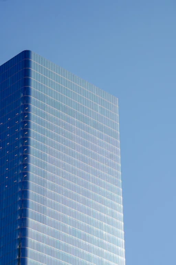 A modern high-rise building with a sleek glass facade reflecting the clear blue sky. The building's structure is composed of large, reflective window panels arranged in a grid, creating a minimalist and contemporary architectural design.