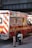 A New York City fire department ambulance is parked on the street. The vehicle is red with white and yellow stripes, featuring the F.D.N.Y logo and markings. A fire hydrant and a metal pole are visible nearby, with an overpass in the background.