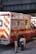 A New York City fire department ambulance is parked on the street. The vehicle is red with white and yellow stripes, featuring the F.D.N.Y logo and markings. A fire hydrant and a metal pole are visible nearby, with an overpass in the background.
