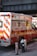 A New York City fire department ambulance is parked on the street. The vehicle is red with white and yellow stripes, featuring the F.D.N.Y logo and markings. A fire hydrant and a metal pole are visible nearby, with an overpass in the background.