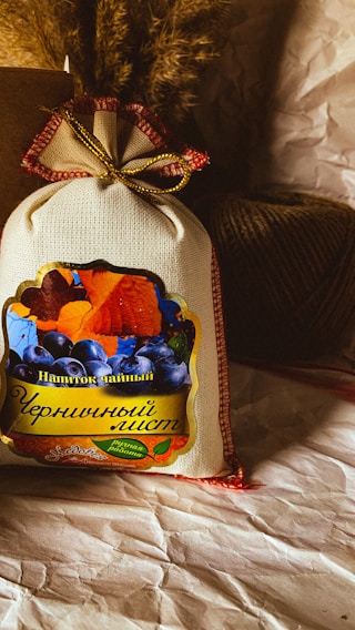 A rustic, textured surface highlights a small burlap sack with a colorful label featuring images of berries and autumn leaves. A box and a ball of twine are positioned in the background, alongside some dried plants.
