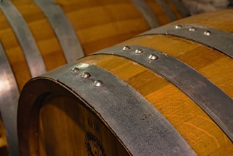A close-up view of wooden barrels with metal bands fastened by bolts. The wood has a rich, warm tone, and the metal appears weathered, indicating use in a cellar or storage setting.