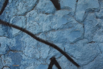 A textured blue concrete wall with dark diagonal spray-painted lines. The surface shows signs of wear with cracks and uneven coloring, giving an urban, gritty aesthetic.