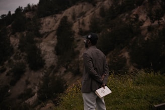 A person stands on a grassy area holding a book behind their back. The individual is wearing a dark jacket and cap, facing a backdrop of rugged terrain covered with scattered trees, suggesting a natural outdoor setting.