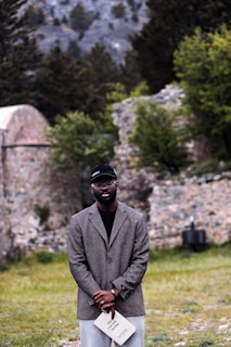 A person stands in an outdoor setting surrounded by greenery and stone structures. They are wearing a dark jacket and cap, and holding a book.