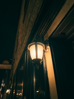 An outdoor wall lantern illuminates a dimly lit alley or street with a warm glow, casting shadows on the brick wall. The lighting creates a cozy and somewhat mysterious atmosphere with other buildings faintly visible in the background.
