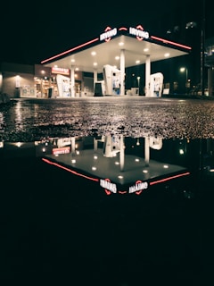 A gas station at night with bright lights illuminating the area. The station is reflected in a pool of water on the asphalt, creating a mirror-like effect. The surroundings are dimly lit, emphasizing the contrast between the bright station and the dark night.