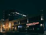 A nighttime urban scene featuring a well-lit restaurant and sports bar with the name 'Boston Pizza' displayed prominently in red neon lights. The building appears modern, with a large office structure in the background. Streetlights cast a warm glow on the wet pavement, suggesting recent rain.