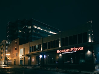 A nighttime urban scene featuring a well-lit restaurant and sports bar with the name 'Boston Pizza' displayed prominently in red neon lights. The building appears modern, with a large office structure in the background. Streetlights cast a warm glow on the wet pavement, suggesting recent rain.